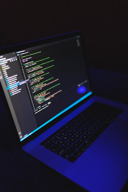 A close-up image of a laptop computer displaying a dark-themed code editor with multicolored lines of programming code. The laptop is positioned on a surface in a dimly lit environment, with the scree