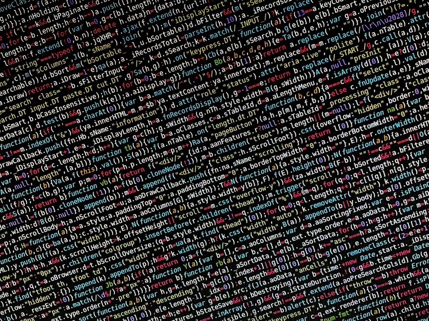 A close-up image of a laptop computer displaying a dark-themed code editor with multicolored lines of programming code. The laptop is positioned on a surface in a dimly lit environment, with the scree